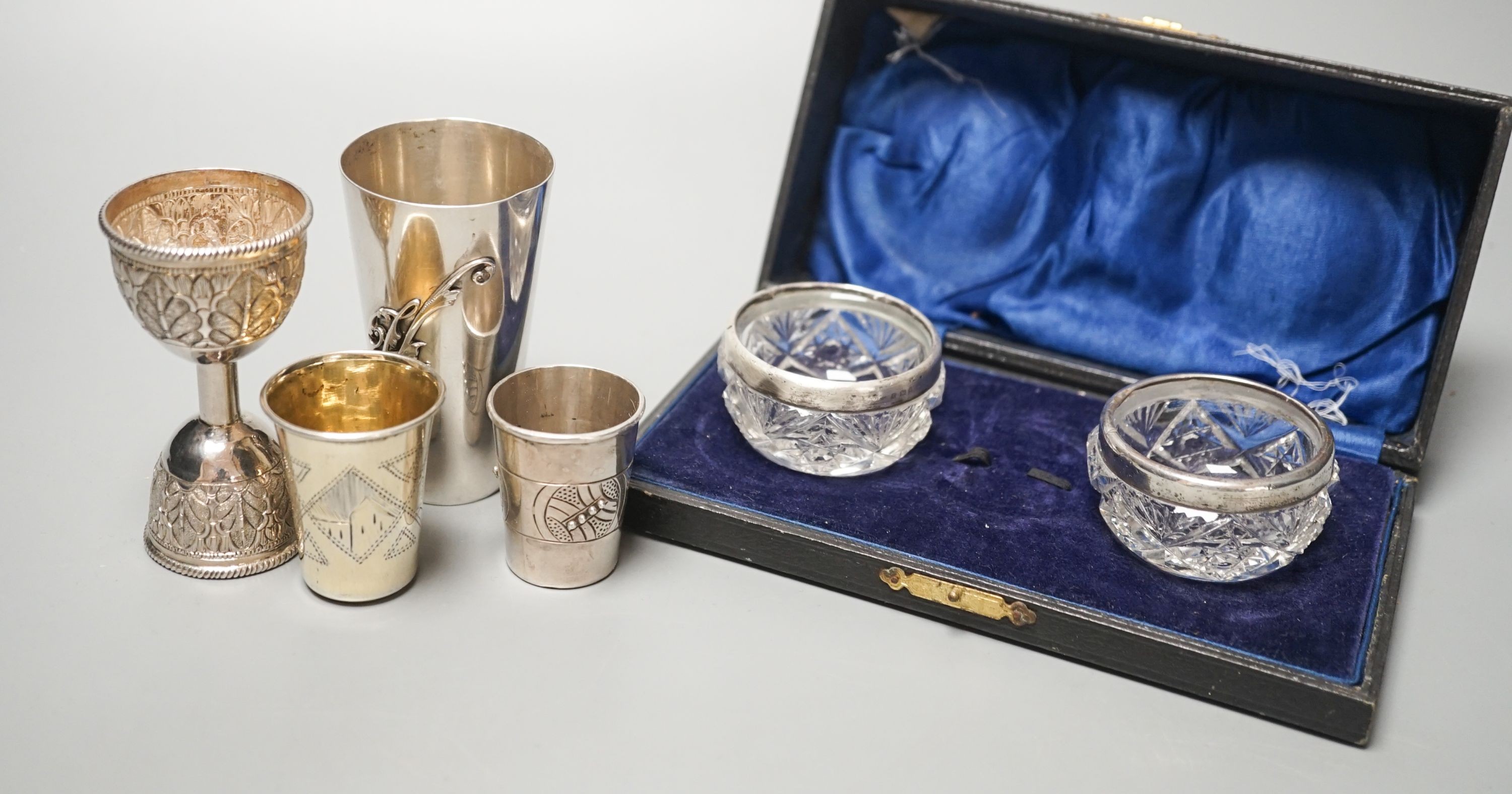 Two early 20th century Russian white metal tots, a sterling beaker, white metal double measure and a case pair of silver mounted glass salts.